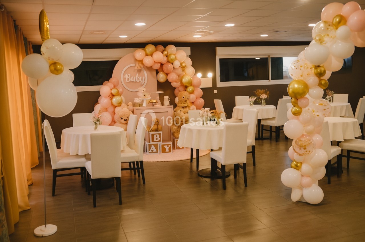Salle décorée pour une fête avec des ballons roses et dorés, des tables dressées et un espace central thématique pour bébé.