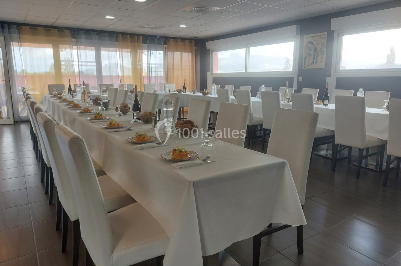 Salle de restaurant lumineuse avec de longues tables dressées, nappes blanches et chaises rembourrées.