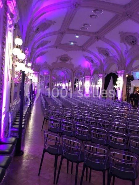 Salle de réception avec plafond orné, éclairage violet et rangées de chaises transparentes alignées.