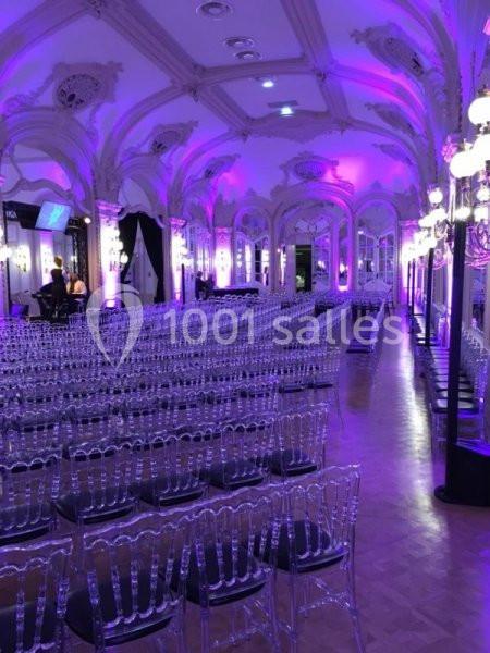 Salle de réception élégante avec des chaises transparentes alignées, éclairée par des lumières violettes et des lustres.