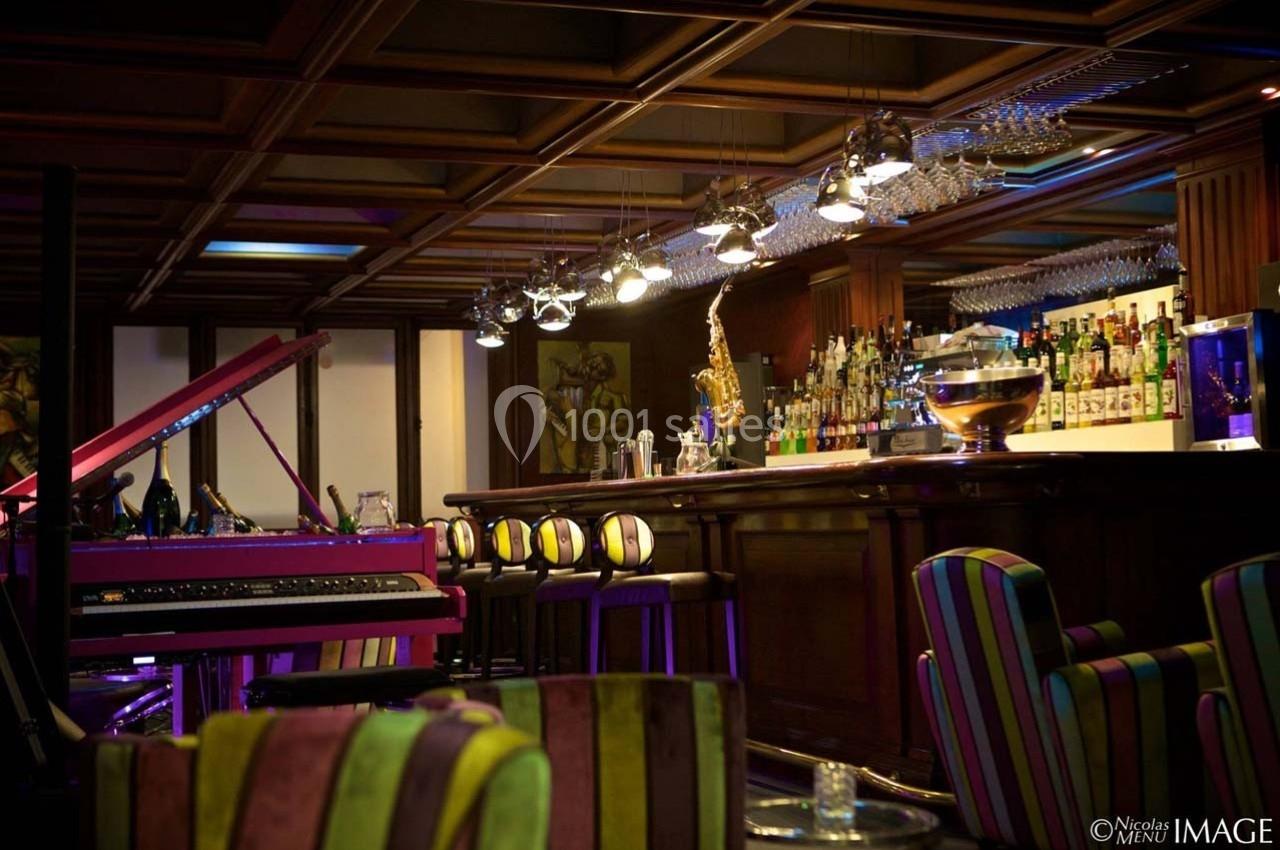 Intérieur d'un bar avec comptoir en bois, tabourets colorés, piano rose et éclairage tamisé.