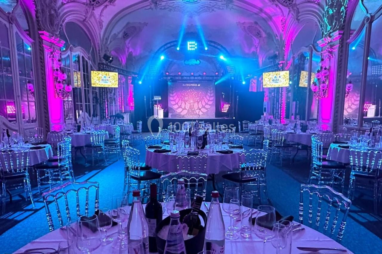 Salle de réception élégante avec tables dressées, éclairage coloré et scène équipée pour un événement.