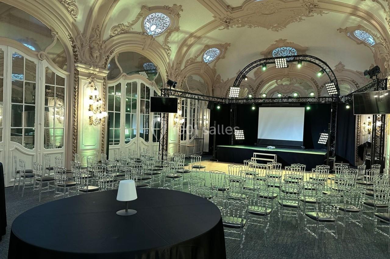 Salle élégante avec décorations baroques, équipée d'une scène, d'éclairages et de chaises transparentes pour un événement.