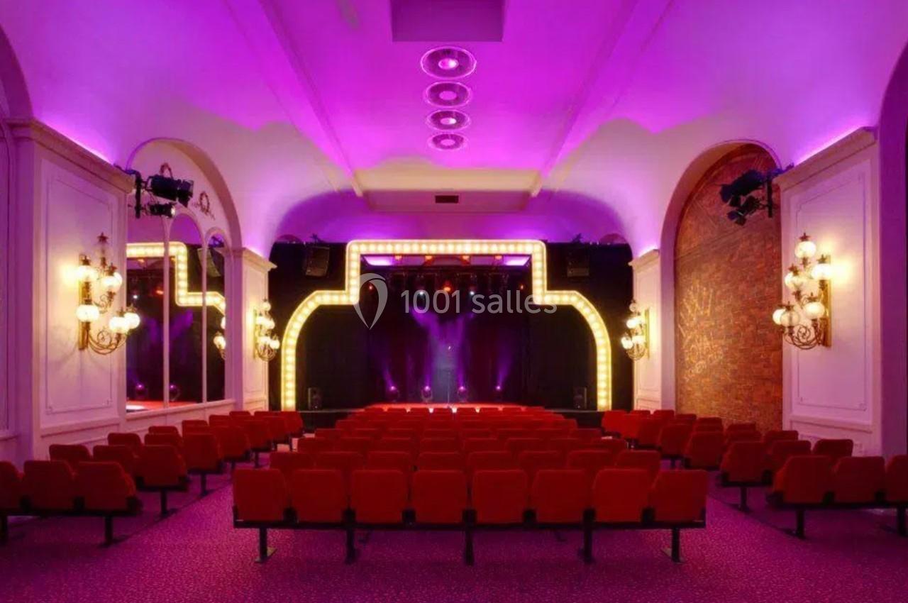 Salle de spectacle avec sièges rouges, scène éclairée par des projecteurs et ambiance lumineuse violette.