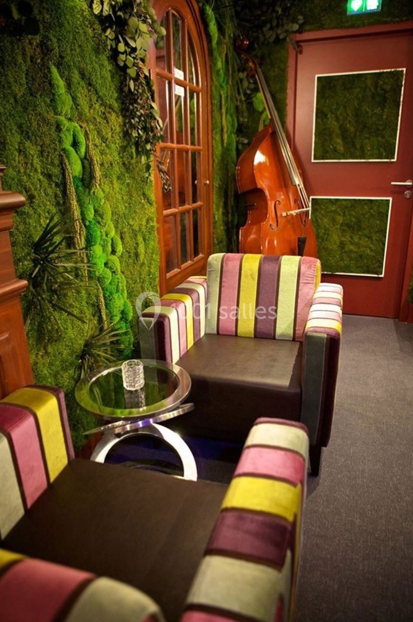 Deux fauteuils colorés et une table en verre dans une pièce avec murs végétalisés, contrebasse et porte rouge.