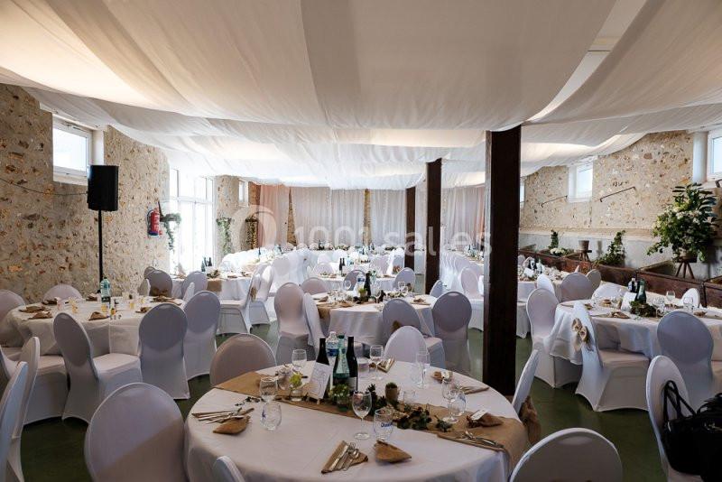 Salle de réception décorée avec des tables rondes, nappes blanches, chaises houssées et plafond drapé de tissu clair.