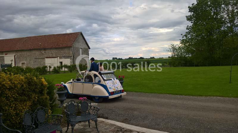 Une voiture ancienne décorée quitte une cour de ferme entourée de verdure sous un ciel nuageux.