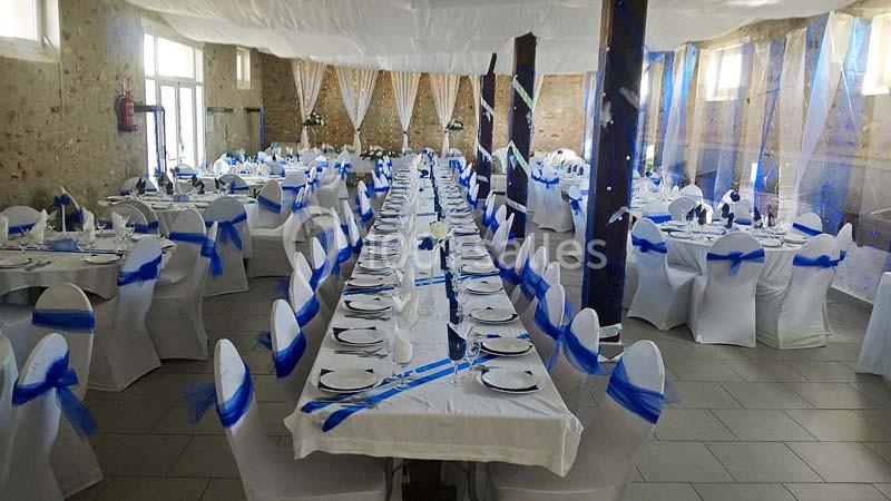 Salle de réception décorée avec des tables alignées, nappes blanches et rubans bleus sur les chaises.