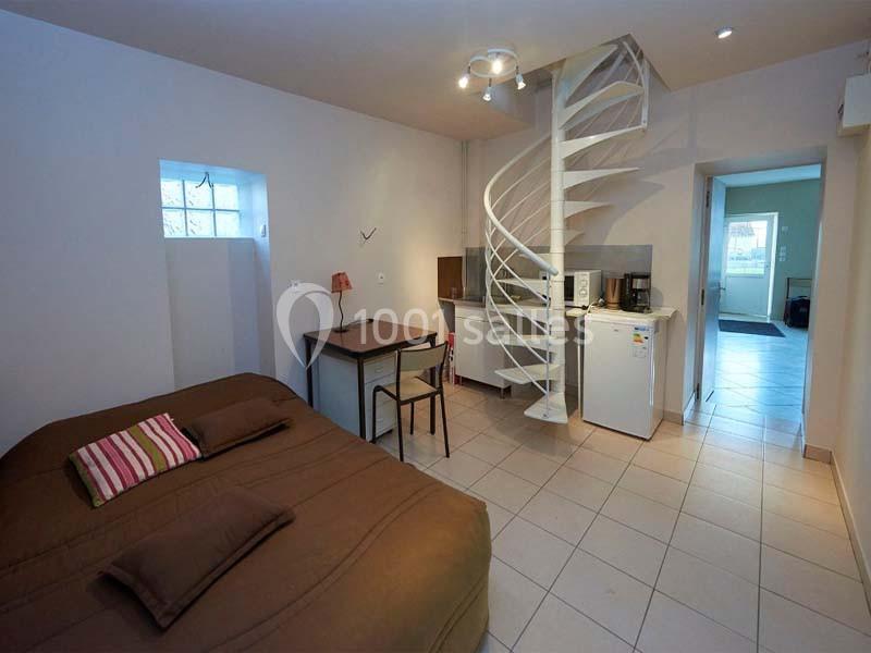 Studio avec lit, table, kitchenette équipée, escalier en colimaçon et carrelage au sol dans un espace lumineux.
