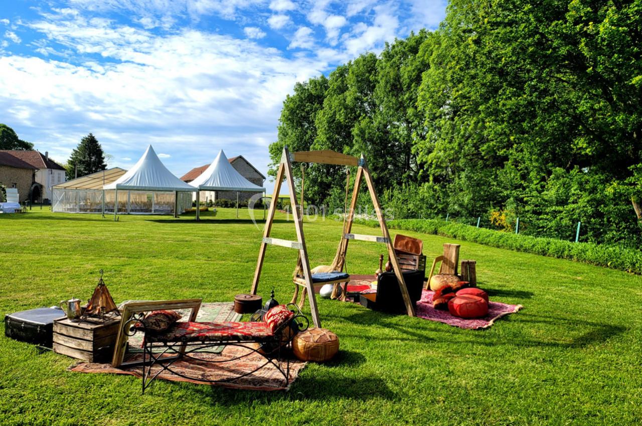 Espace extérieur aménagé avec tapis, coussins et balancelle, entouré de verdure et de tentes blanches au loin.