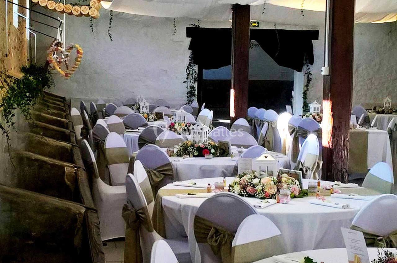 Salle décorée pour un événement, avec des tables rondes ornées de fleurs et des chaises couvertes de housses blanches.