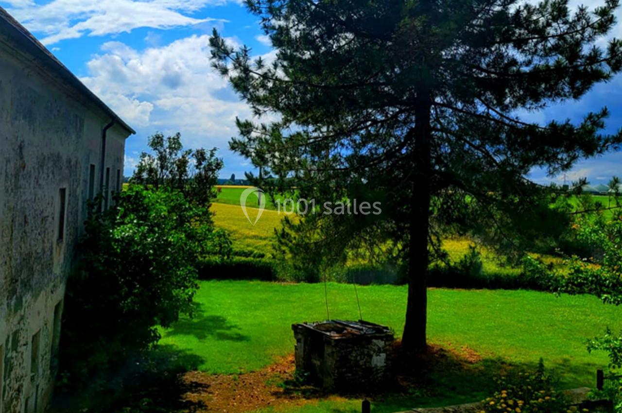 Vue d'un jardin verdoyant avec un puits en pierre, un grand arbre et des champs sous un ciel partiellement nuageux.