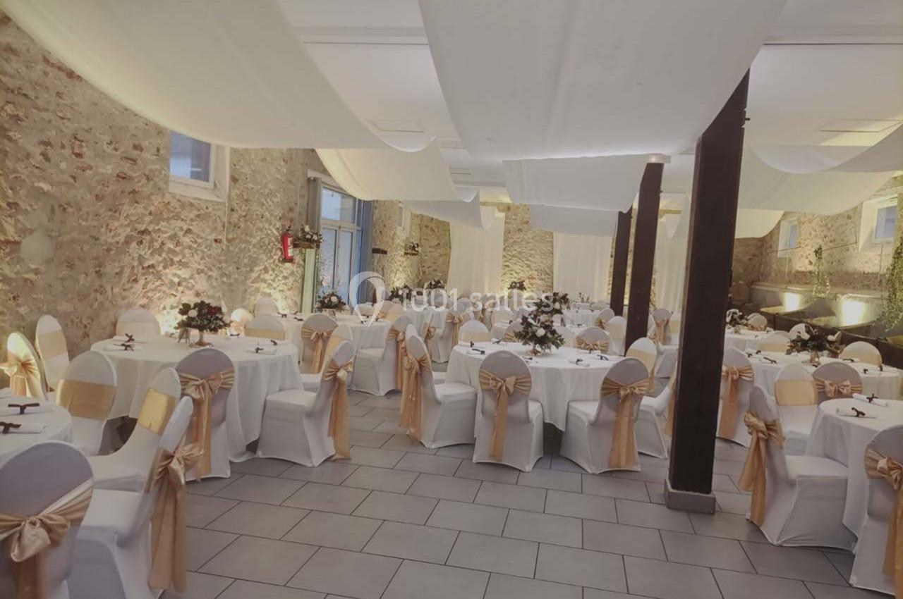 Salle de réception décorée avec des tables rondes, nappes blanches, chaises ornées de rubans dorés et murs en pierre…