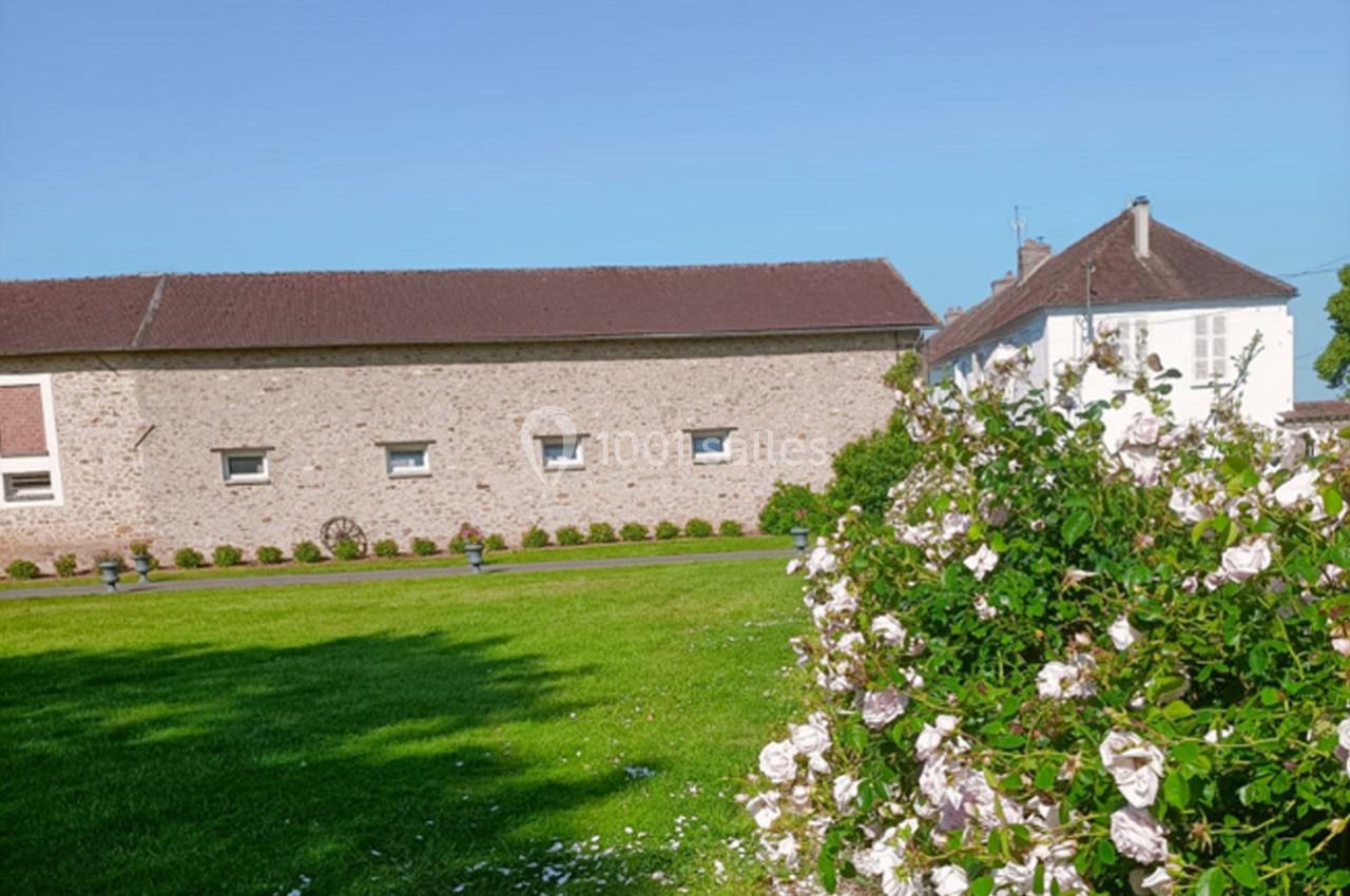 Bâtiment en pierre avec toit en tuiles, pelouse verte et arbuste fleuri sous un ciel bleu clair.