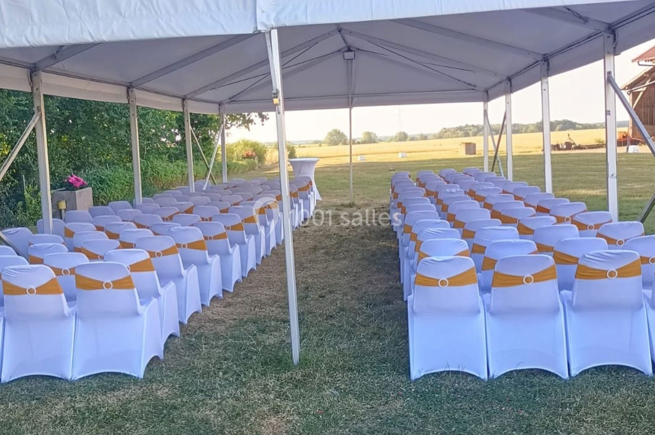 Chaises blanches décorées de rubans jaunes alignées sous une tente en extérieur, avec vue sur un champ.