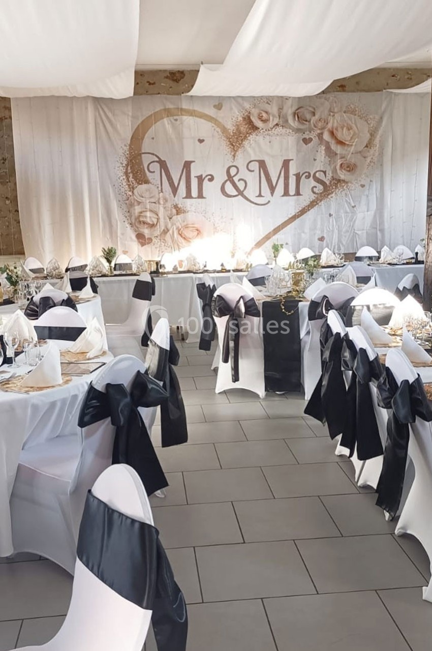 Salle de réception décorée pour un mariage, avec des tables dressées et un fond ’Mr & Mrs’ orné de fleurs.