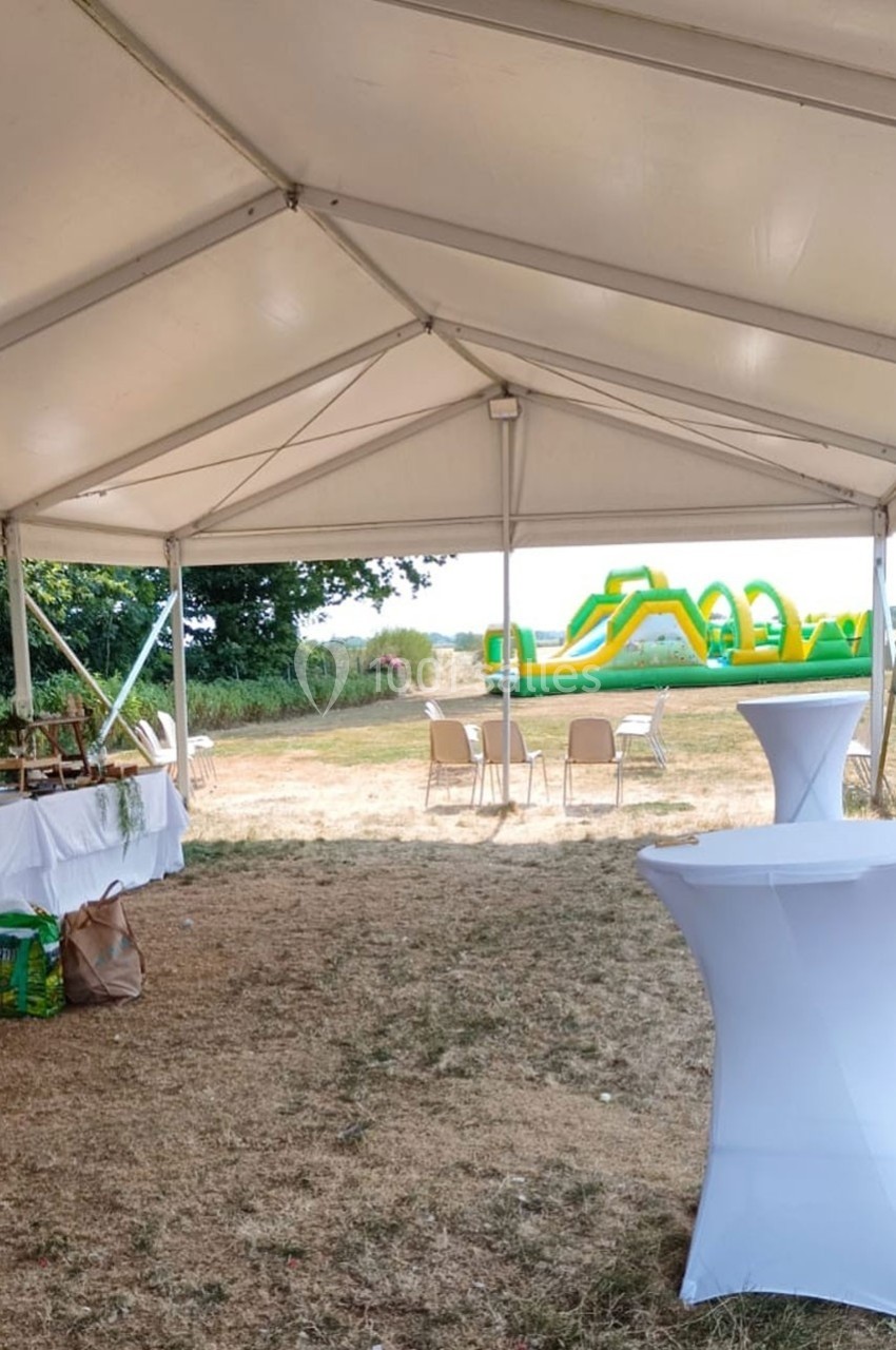 Tente blanche ouverte avec tables hautes et vue sur un parcours gonflable vert dans un espace extérieur.