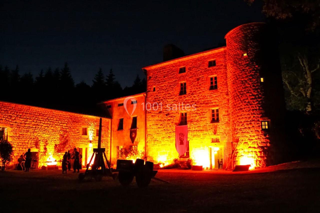 Façade d'un château en pierre illuminée par des lumières rouges et oranges, avec des silhouettes de visiteurs.