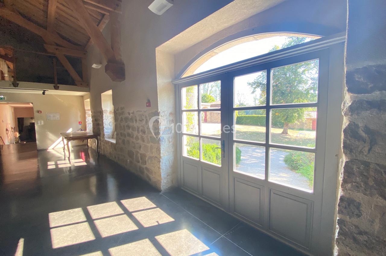 Grande porte vitrée laissant entrer la lumière dans un couloir aux murs en pierre et au plafond en bois.