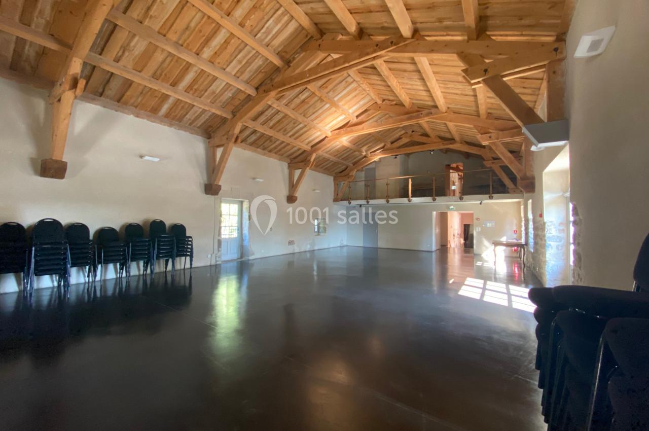 Grande salle avec charpente en bois apparente, sol en béton lisse, chaises empilées sur le côté et lumière naturelle.