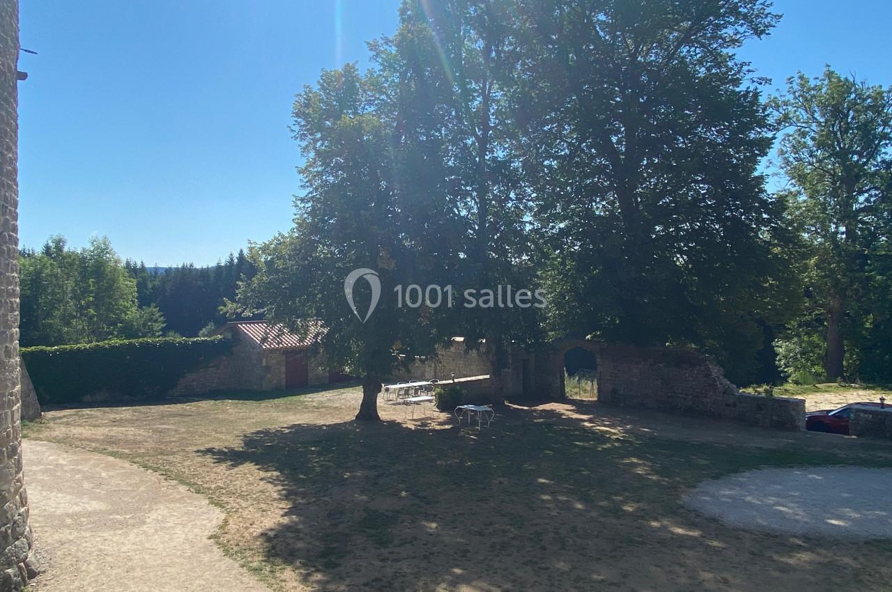 Cour extérieure ensoleillée avec des arbres, un mur en pierre, une table et des chaises à l'ombre.