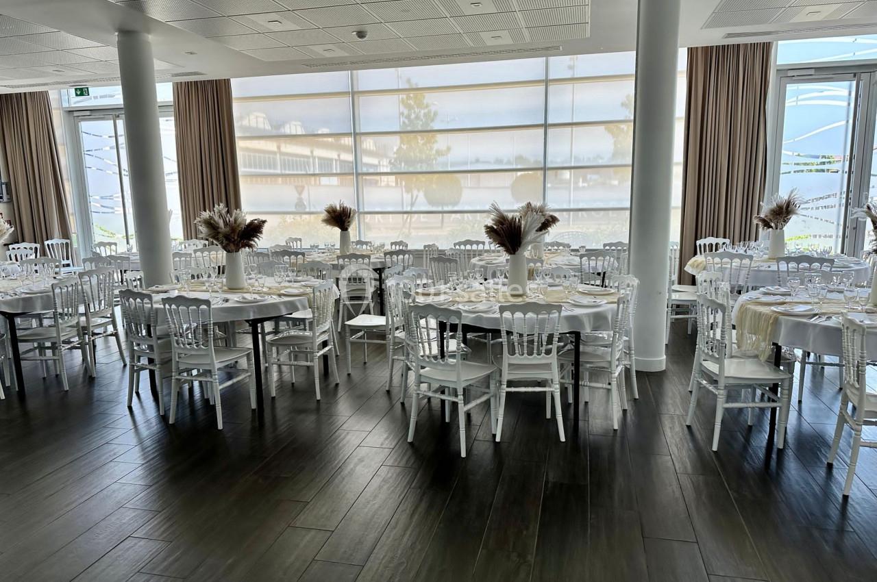 Salle de réception lumineuse avec tables rondes décorées de bouquets de fleurs séchées et chaises blanches.
