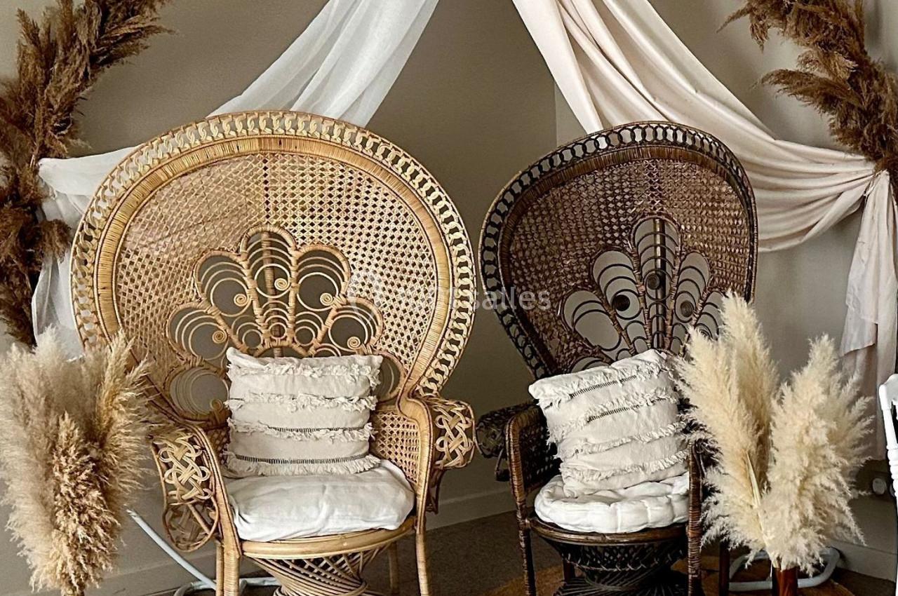 Deux fauteuils en rotin ornés de coussins, disposés sous une arche décorée de voilages et de pampas.