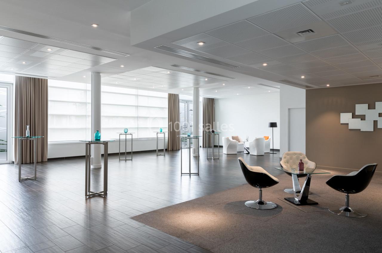 Salle lumineuse avec parquet sombre, mobilier moderne, tables hautes et fauteuils disposés dans un espace ouvert.