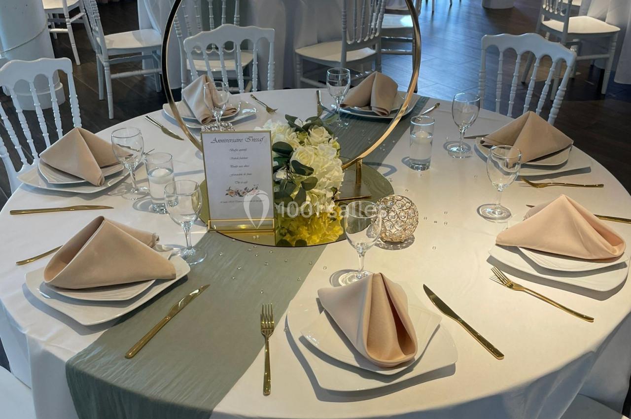 Table ronde élégamment dressée avec nappes blanches, serviettes beige pliées, décoration florale et couverts dorés.