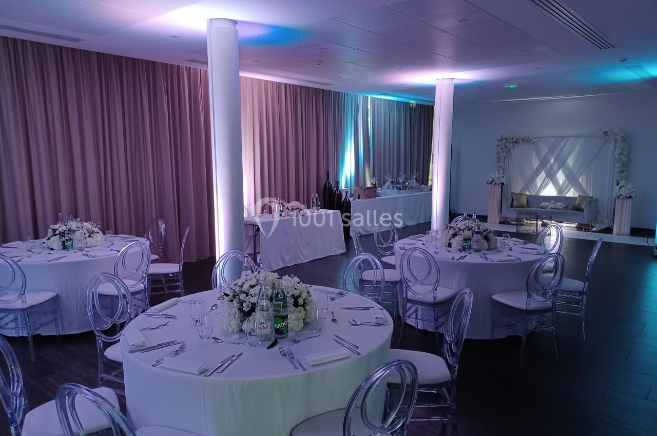 Salle de réception élégante avec tables rondes dressées, chaises transparentes et éclairage tamisé.