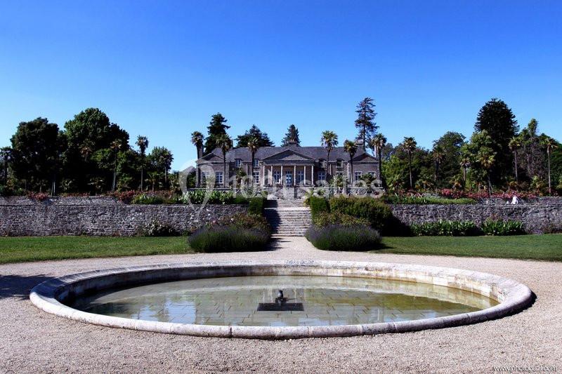 Manoir en pierre entouré de jardins, avec une fontaine circulaire au premier plan sous un ciel dégagé.