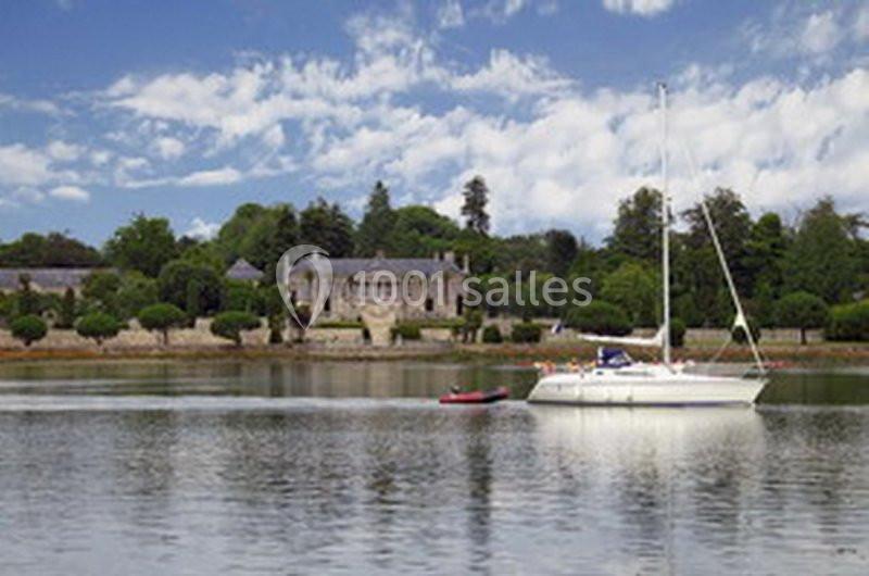Voilier blanc ancré sur une rivière calme, avec un manoir en pierre et des arbres en arrière-plan.