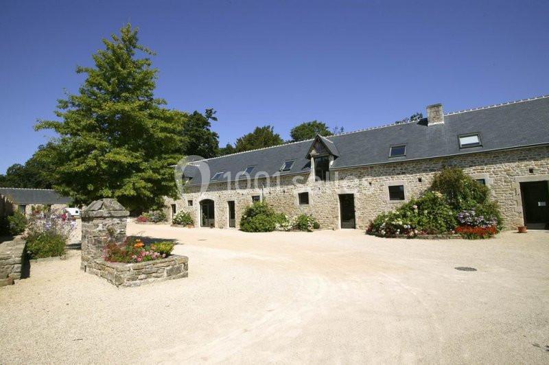 Cour d'une bâtisse en pierre avec toit en ardoise, entourée de fleurs et d'arbres sous un ciel dégagé.