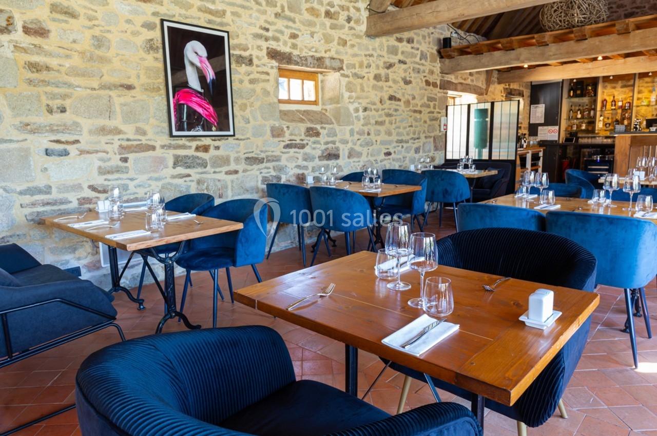 Salle de restaurant avec murs en pierre, tables en bois dressées, chaises bleues et un tableau de flamant rose au mur.