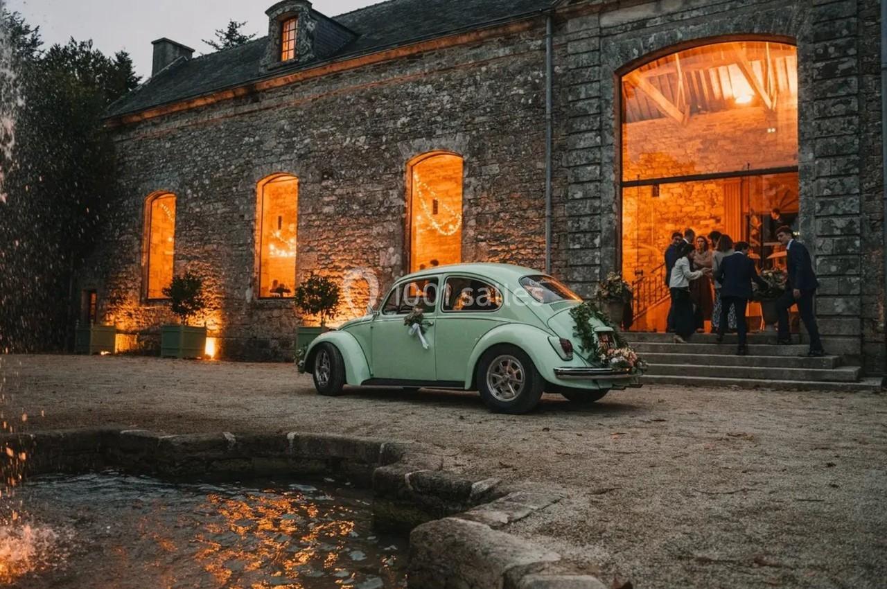 Une voiture ancienne vert clair décorée pour un mariage est garée devant un bâtiment en pierre éclairé.