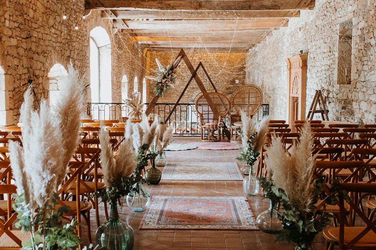 Salle de cérémonie décorée avec des chaises en bois, des herbes de la pampa et une arche triangulaire ornée de fleurs.
