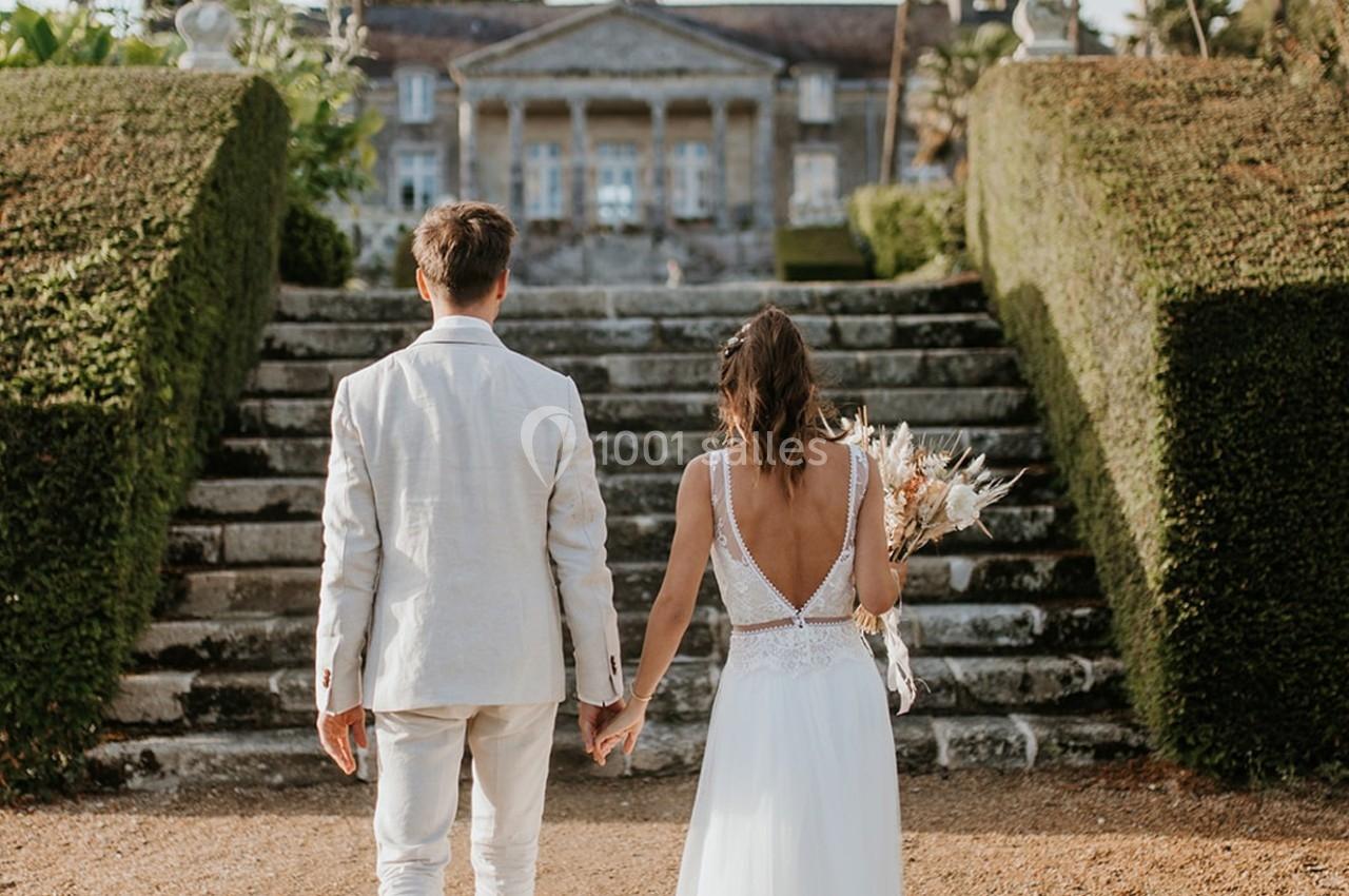 Un couple en tenue de mariage monte des marches en pierre, menant à un bâtiment historique entouré de verdure.