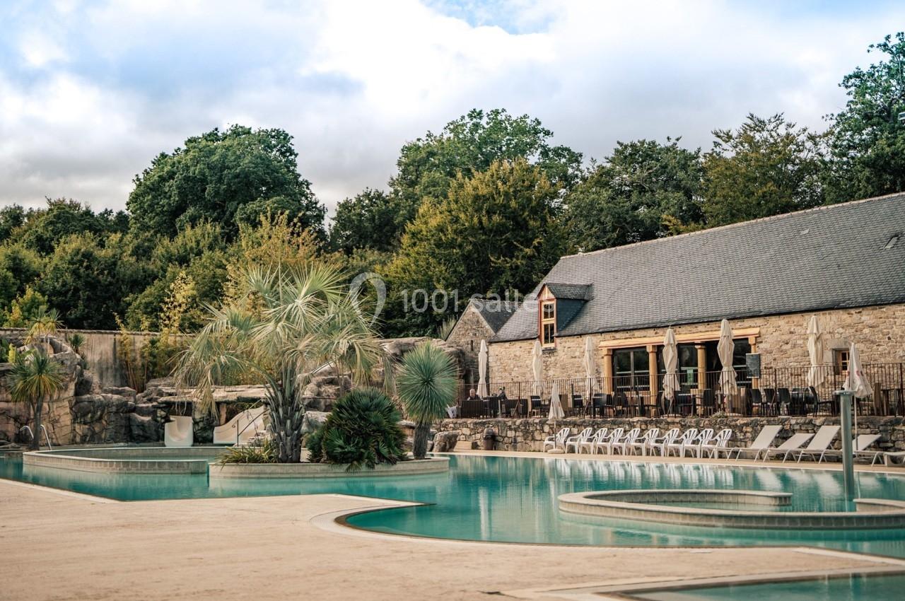 Piscine extérieure avec palmiers, entourée de chaises longues et d'un bâtiment en pierre sous un ciel partiellement nuageux.
