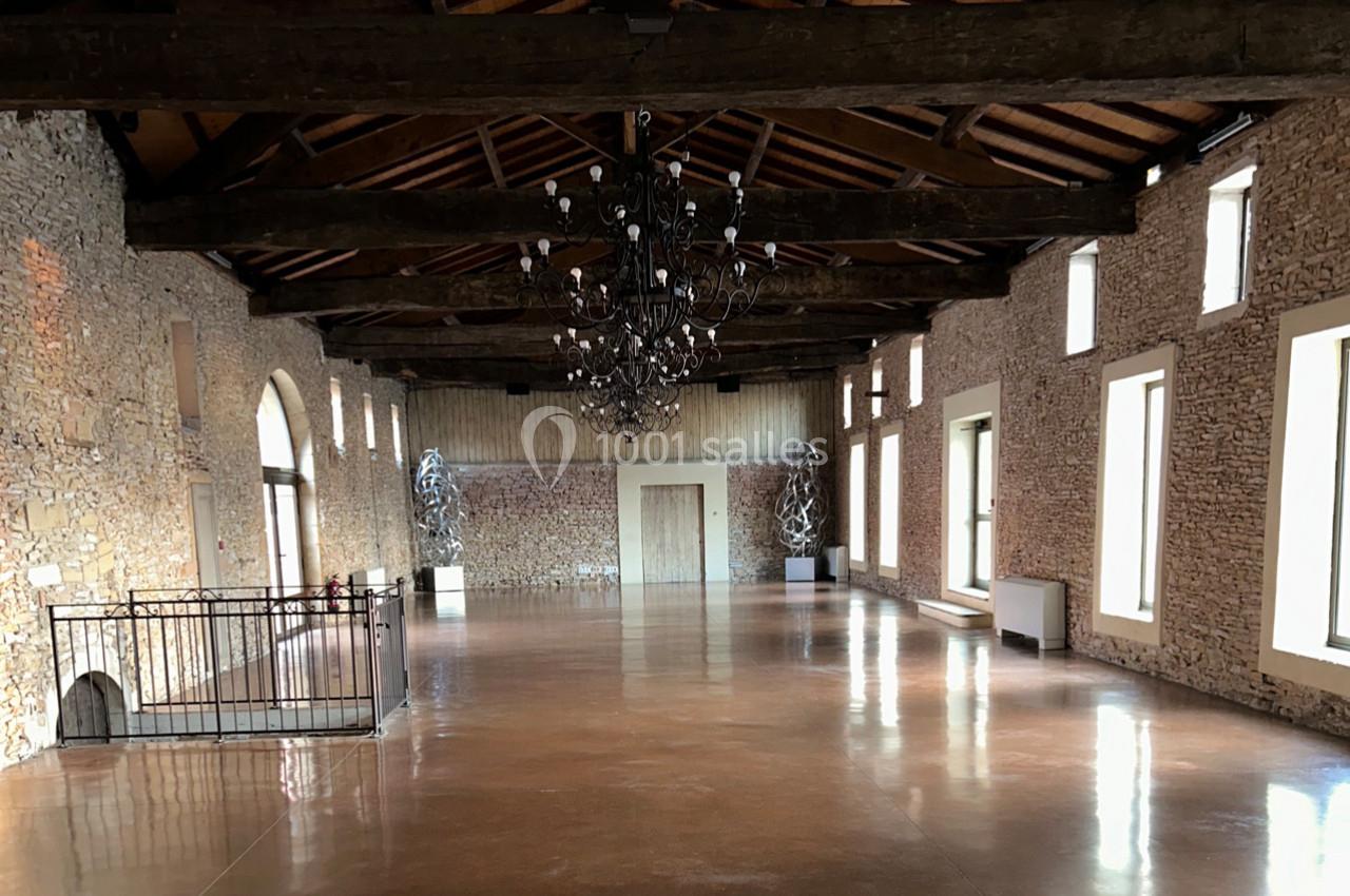 Grande salle en pierre avec plafond en poutres apparentes, lustre central et grandes fenêtres laissant entrer la lumière.