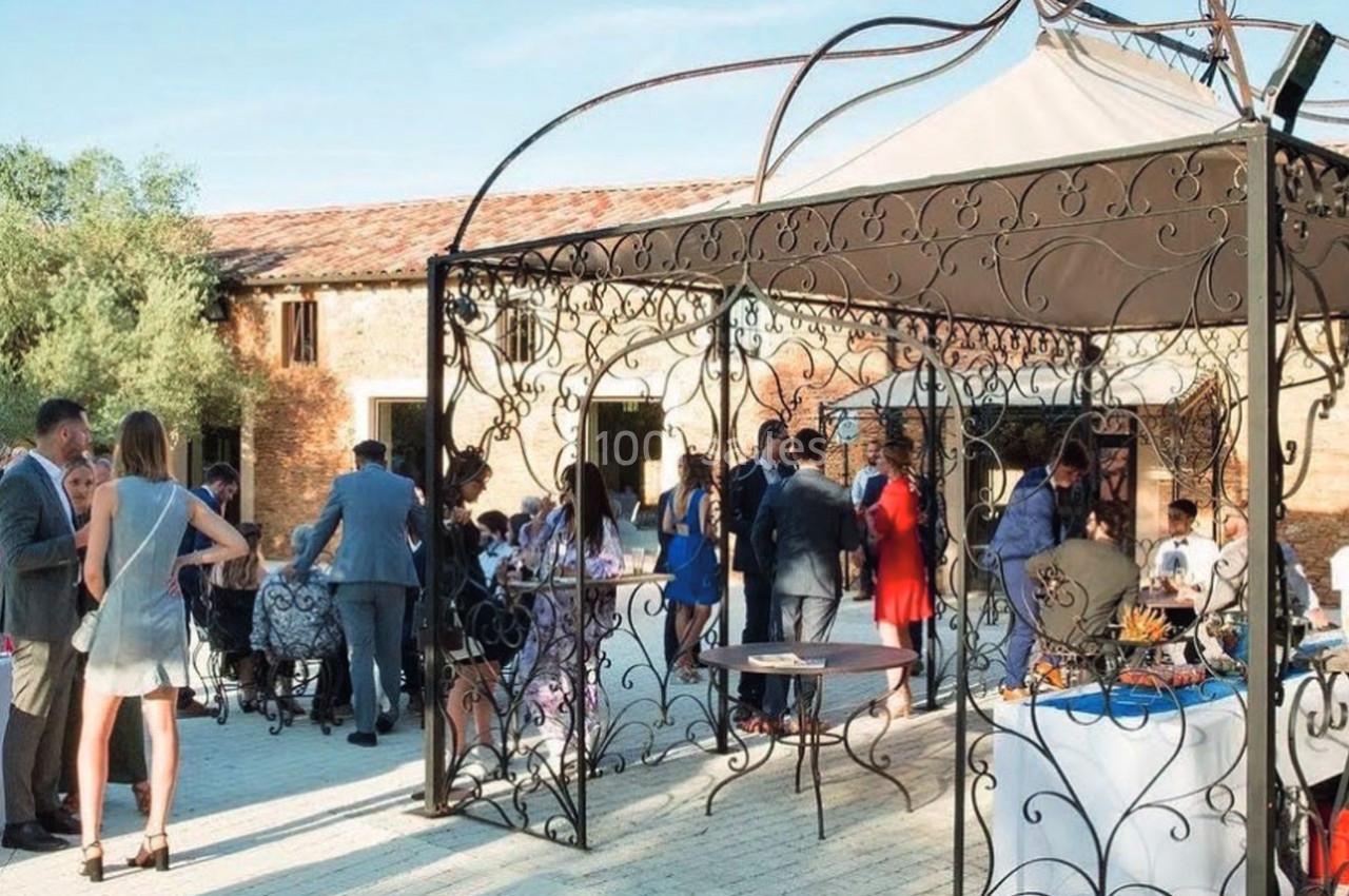 Groupe de personnes rassemblées sous une pergola en fer forgé dans une cour pavée, près d'un bâtiment en pierre.