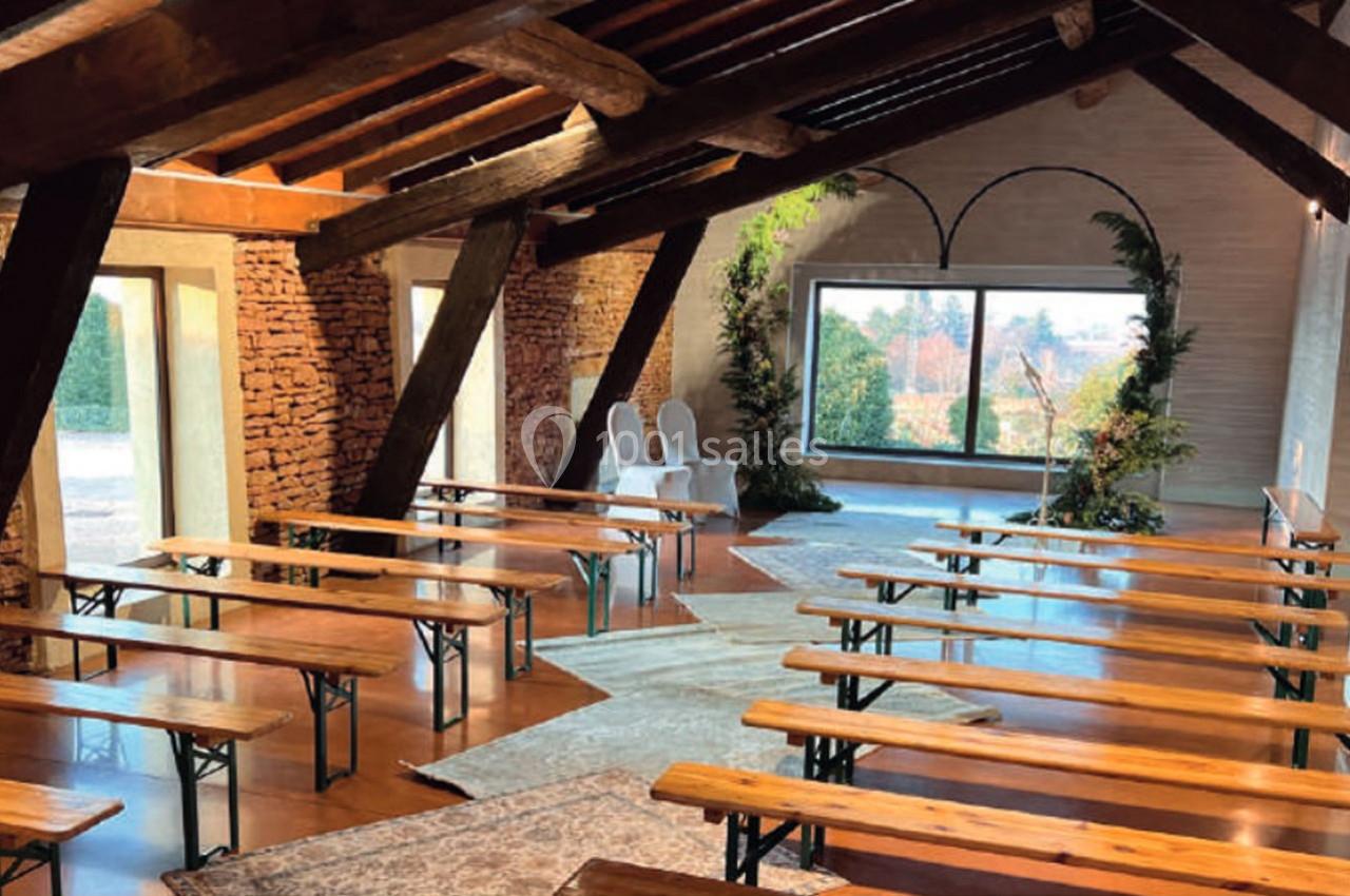 Salle lumineuse avec poutres apparentes, bancs en bois alignés et grande baie vitrée donnant sur un paysage arboré.
