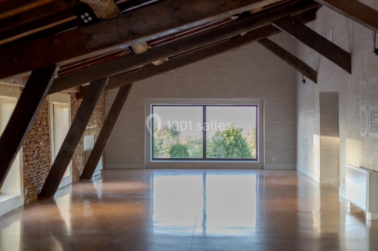 Intérieur d'une pièce mansardée lumineuse avec poutres apparentes, murs clairs et grande fenêtre donnant sur des arbres.