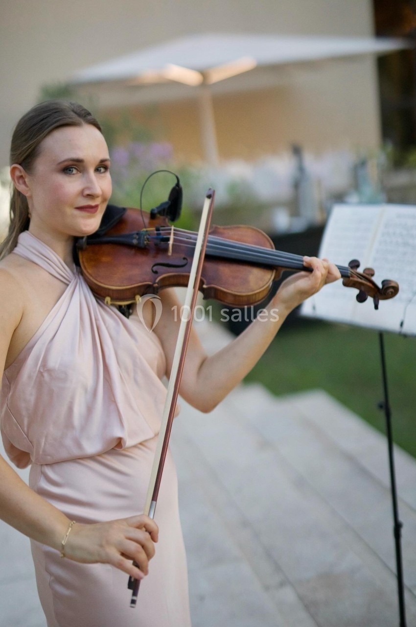 Une musicienne en robe rose joue du violon en extérieur, avec une partition sur un pupitre en arrière-plan.