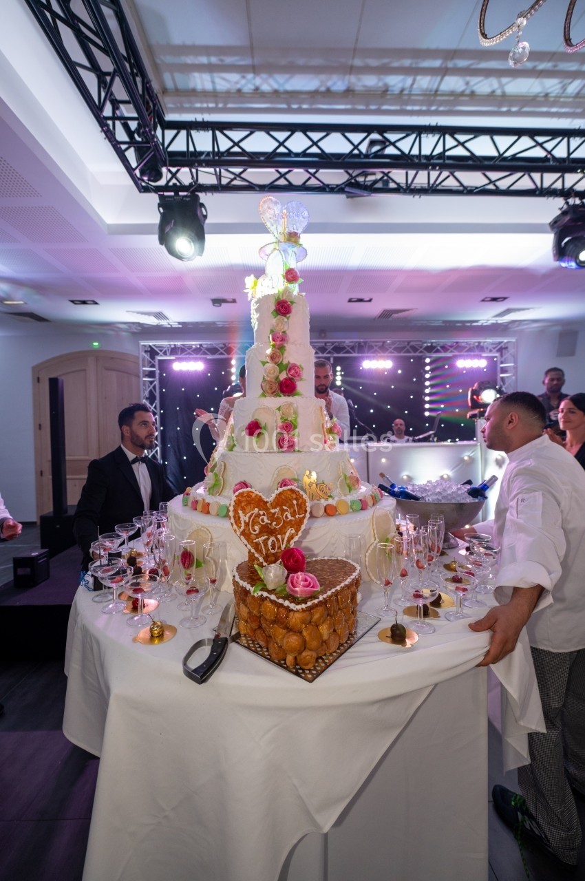 Un gâteau de mariage décoré de fleurs et de figurines, entouré de desserts et de convives dans une salle éclairée.