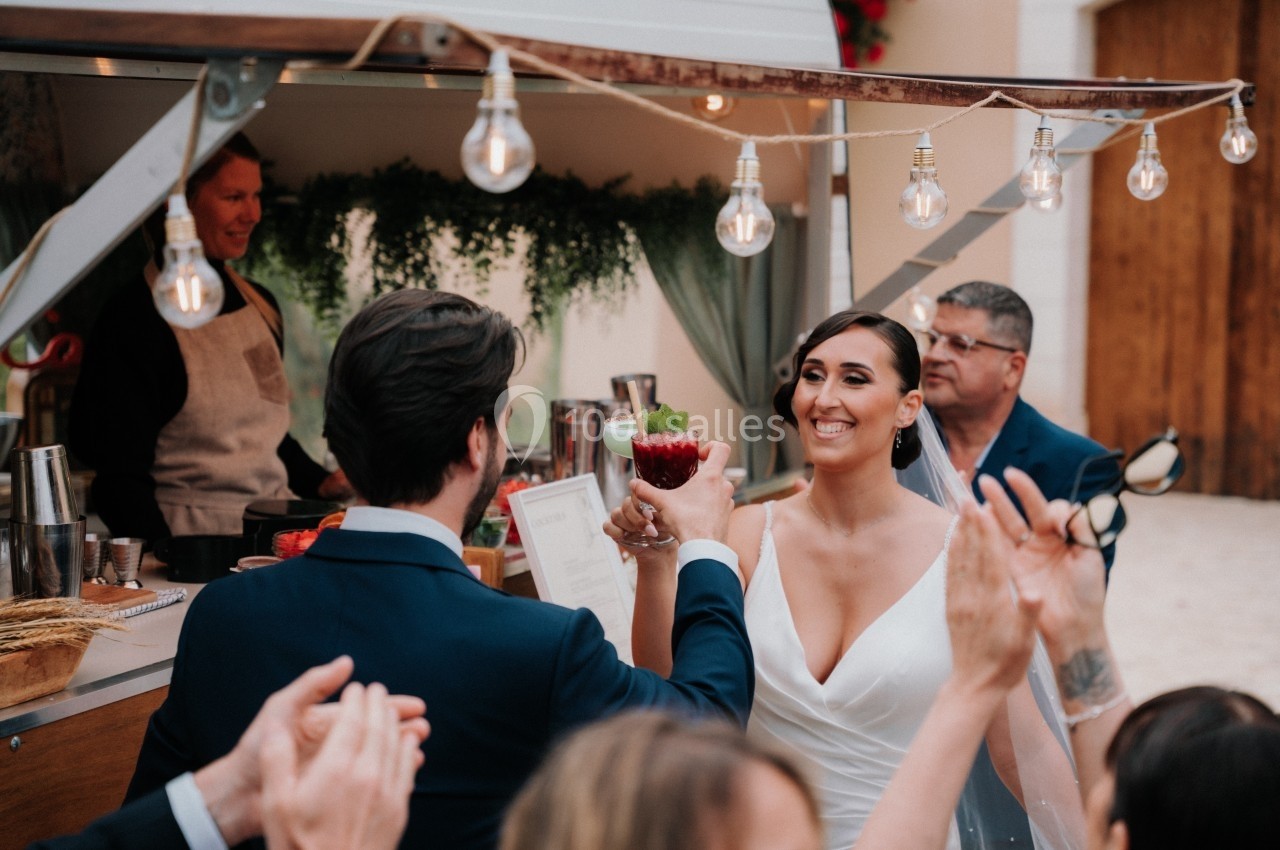 Un couple trinque joyeusement devant un bar mobile décoré, entouré d'invités applaudissant.