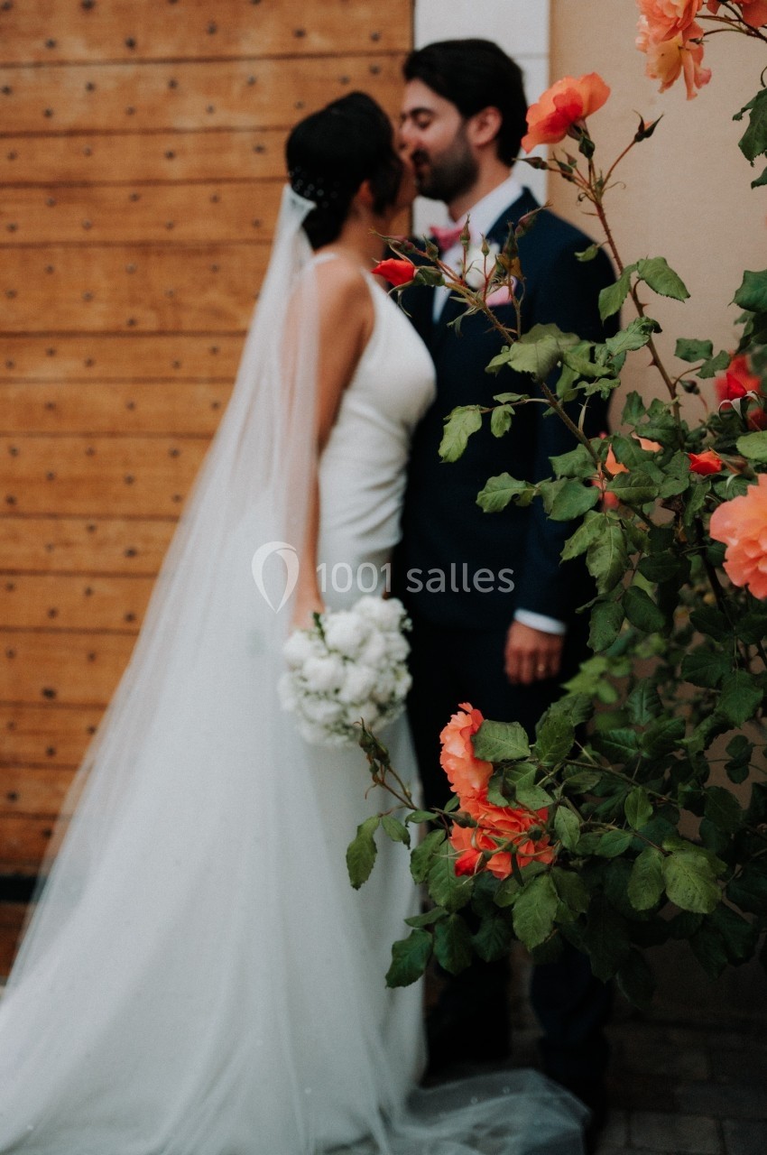 Un couple de mariés s'embrasse devant une porte en bois, avec des fleurs orangées au premier plan.