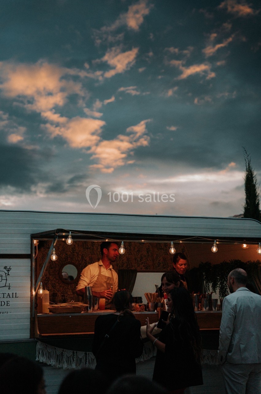 Un bar mobile éclairé par des guirlandes lumineuses, avec des serveurs préparant des boissons pour des clients en soirée.