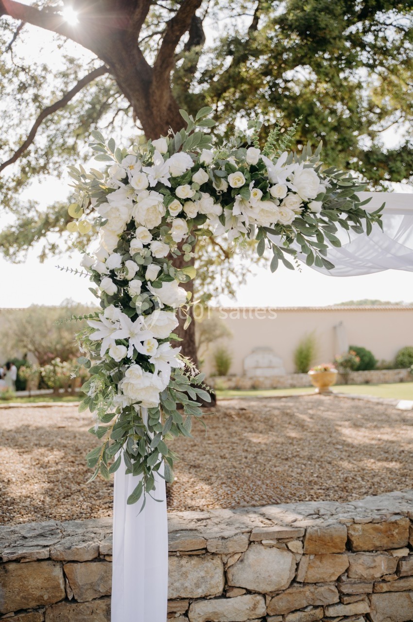 Décoration florale blanche et verte en arc suspendue à une structure, avec un jardin en arrière-plan.