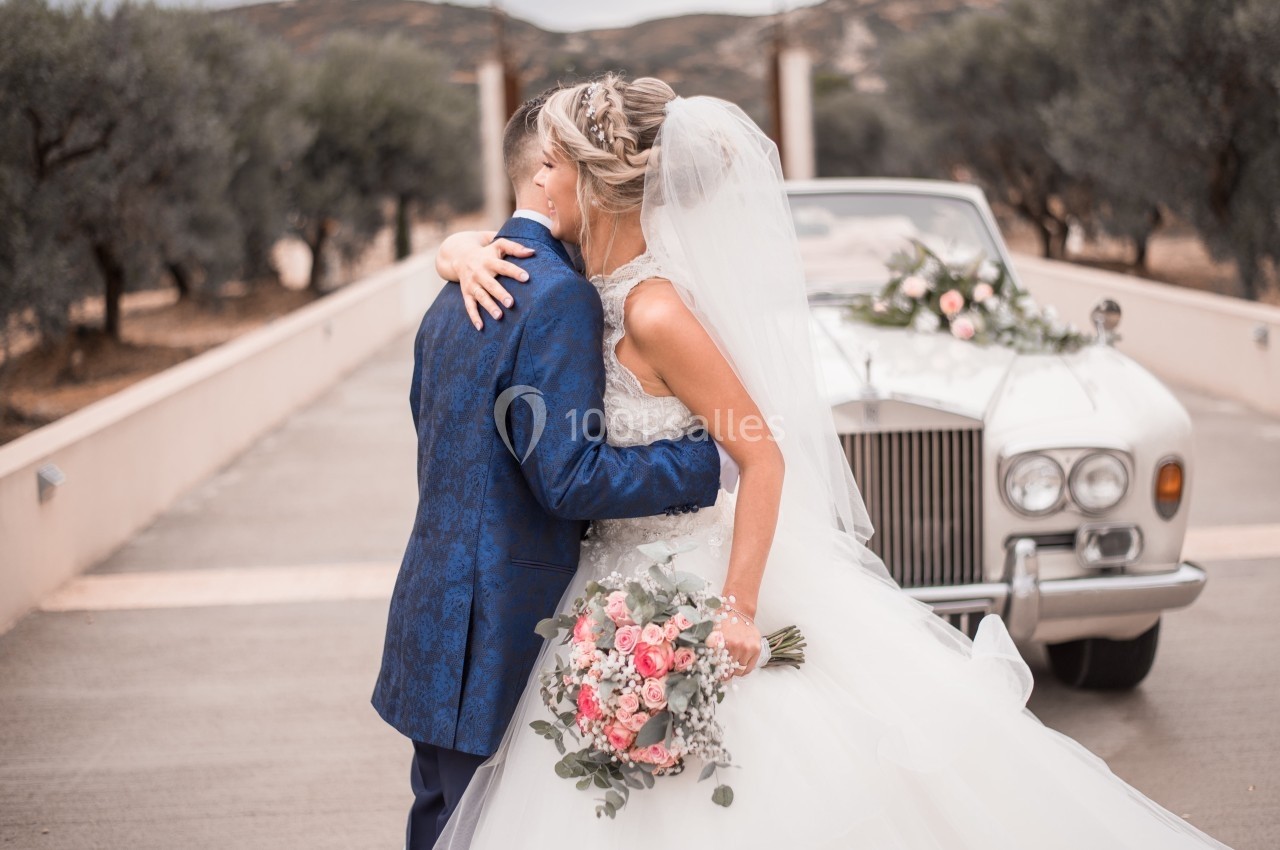 Une mariée en robe blanche et un homme en costume bleu s'enlacent devant une voiture ancienne décorée de fleurs.
