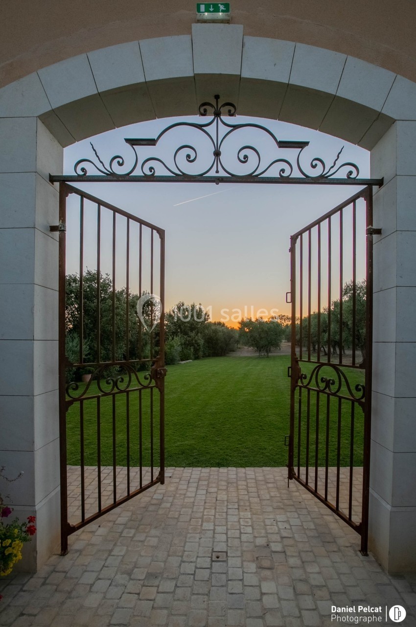 Portail en fer forgé ouvert sur un jardin verdoyant au coucher du soleil, encadré par des oliviers.
